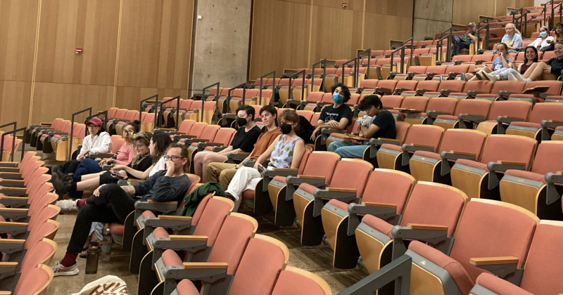 A group of masked and unmasked graduate and undergraduate students sit in a classroom auditorium, waiting for a film to be screened.